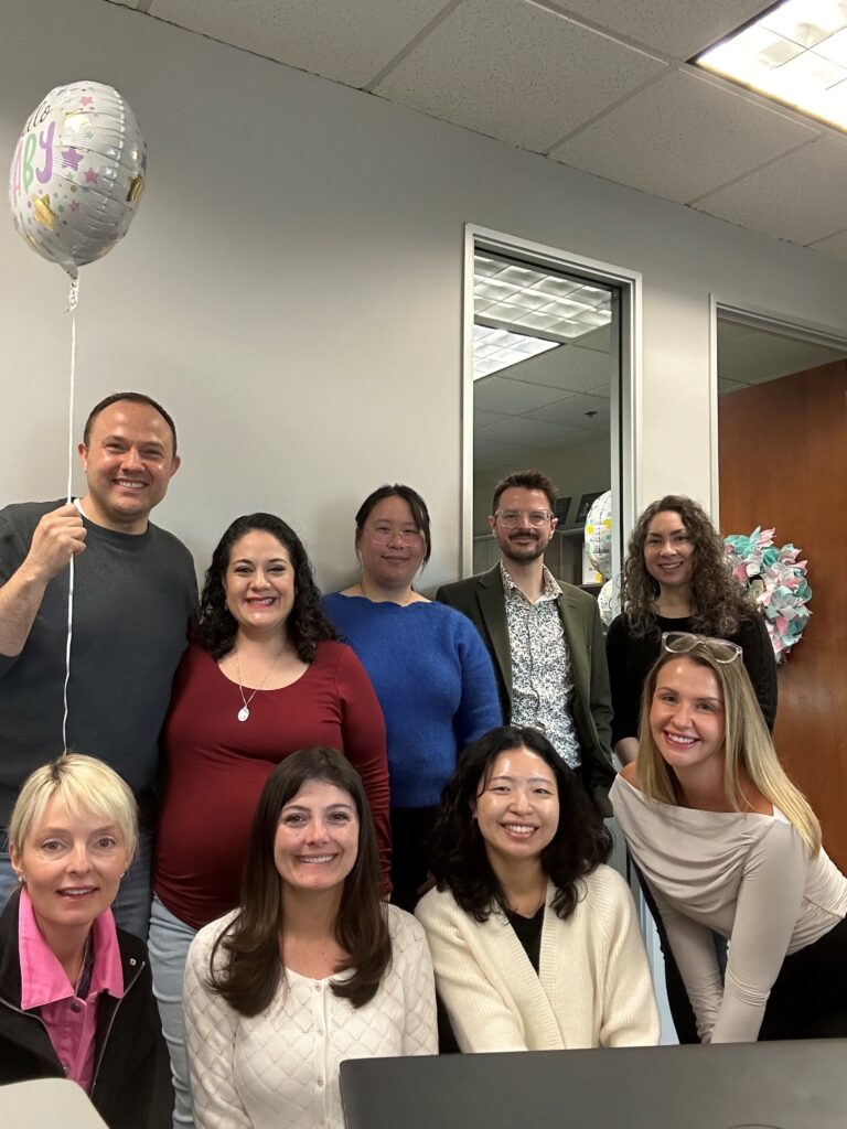 A group of smiling EVALCORP team members gathered in the office to celebrate a colleague's baby shower. One person is holding a balloon that says "Hello Baby,"