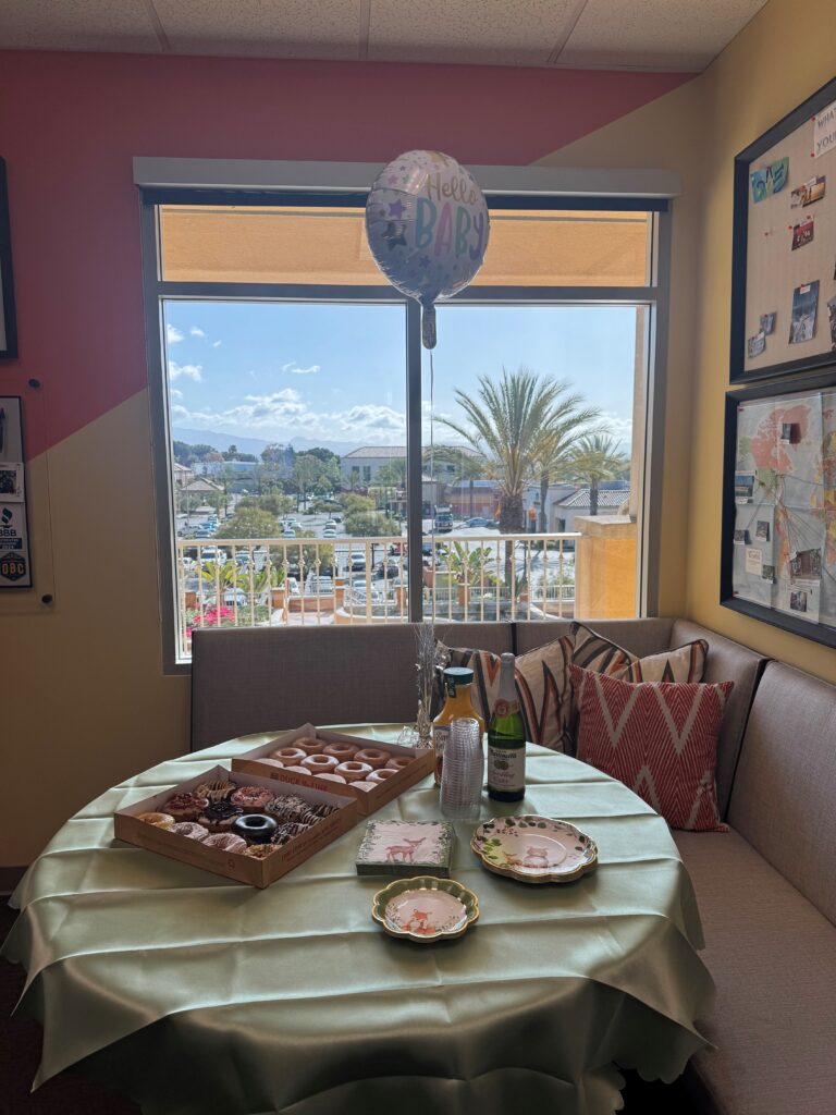 EVALCORP’s office baby shower setup, featuring donuts, celebratory drinks, woodland-themed plates, and a “Hello Baby” balloon. The table is set in a bright corner with a scenic view outside the window.