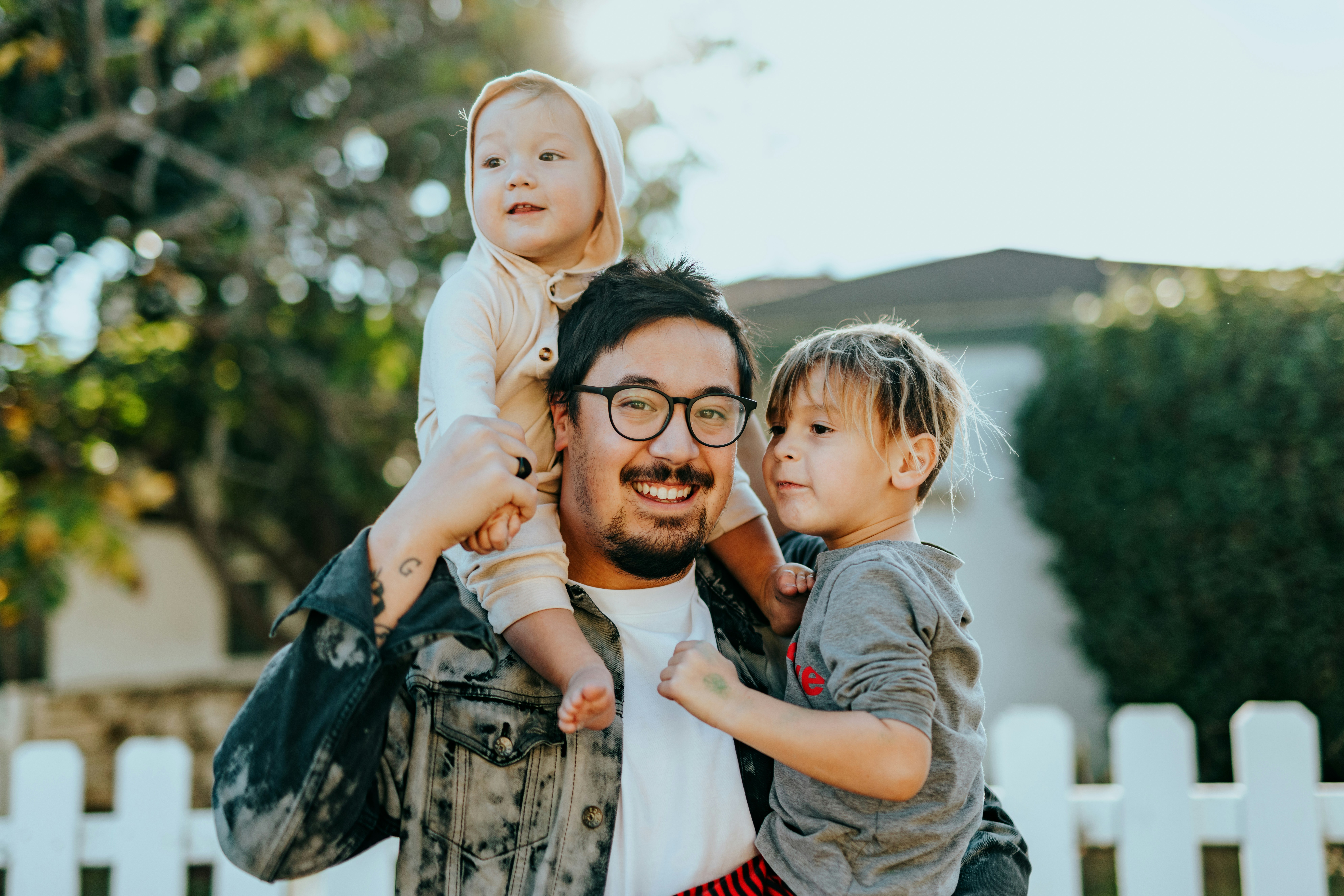 A smiling man holds his two children—his younger child sits on his shoulders while he carries the older child in his arms.