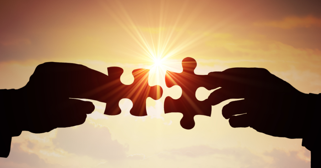 Silhouette of two hands about to join two pieces of a jigsaw puzzle in front of a clouded sky and a brilliant source of light