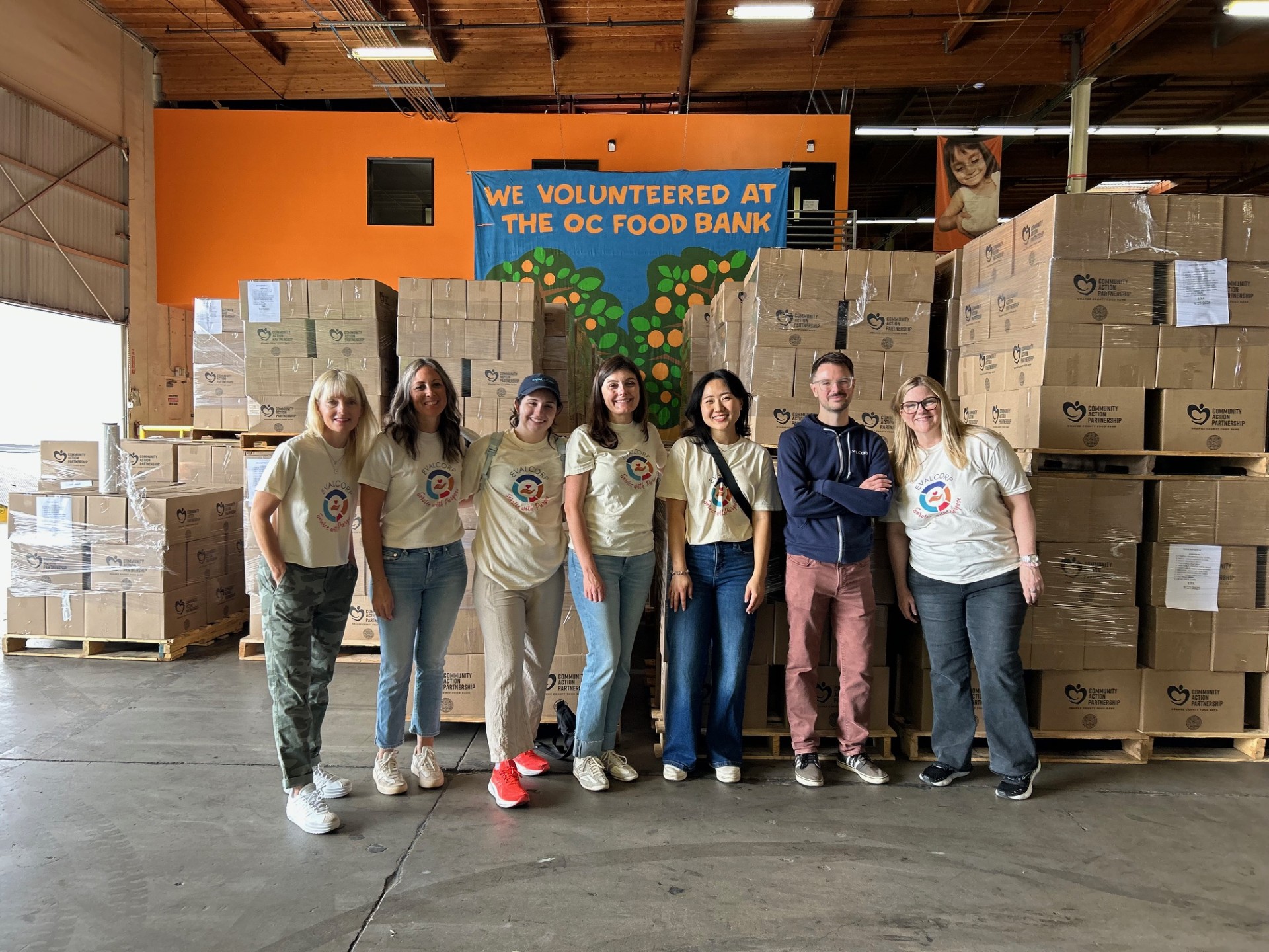Picture of EVALCORP team members in posing in front of boxes they packed at the Orange County food bank. Team members are wearing matching t-shirts that say "Service With Purpose"