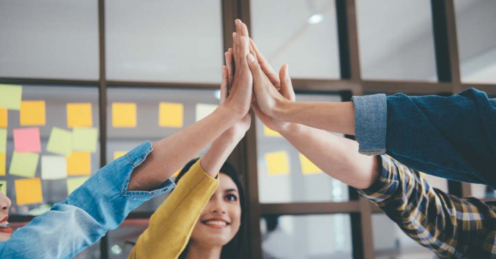 Four people in an implied casual work setting circled up and each putting a hand in the air and touching each other's hands in an implied cheer. The background contains a windowed partition with numerous multicolored sticky notes attached