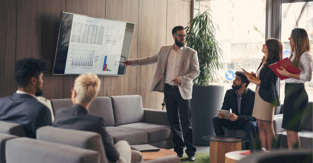 Person giving a presentation referring to chart on a flat screen monitor mounted on a brown wood-grained wall with several people variously standing or sitting in couches and paying attention and/ or taking notes