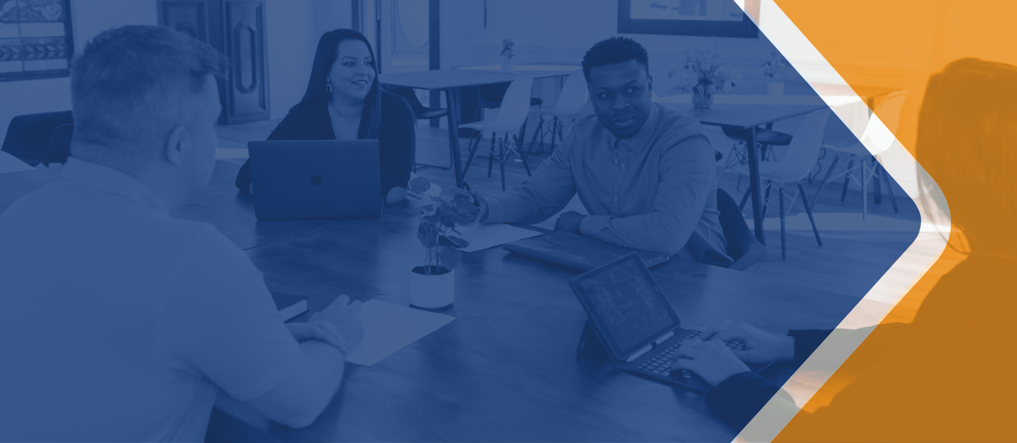 Photograph of people in business casual clothing sitting around a table with laptop computers and sheets of paper in a room with two empty tables and chairs in the background. Overlain over this image is a large area of transparent blue and a smaller area of transparent orange