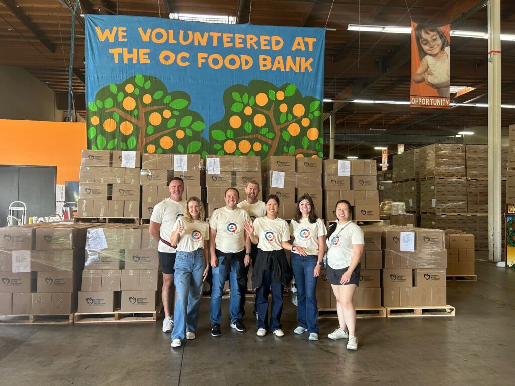 Photograph of EVALCORP team members in t-shirts with "Service with Purpose" logo standing in front of boxes packed for Orange County food pantry.