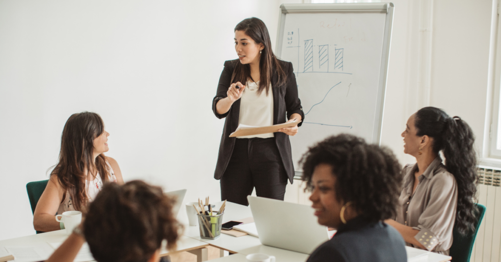 photograph of a woman standing in front of a whiteboard with graphs drawn on it and surrounded by other women sitting around a table