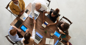 People sitting around a wood table with laptop computers, taking notes and collaborating, and viewed from above the table